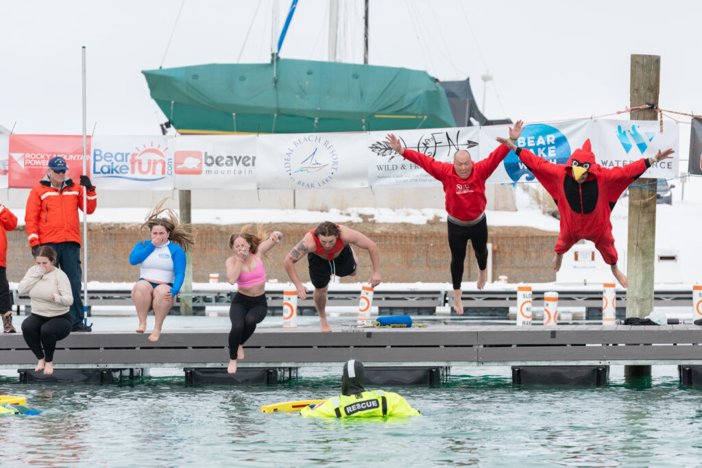 Six people jumping off the dock into Bear Lake in the winter.  One of the people is wearing a bird costume.